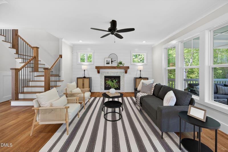Bright living room with wooden staircase, central fireplace, gray sofa, two armchairs on striped rug, and large windows with green views.