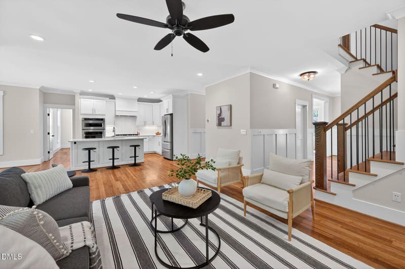 Open-concept living room and kitchen with white cabinets, hardwood floors, striped rug, black ceiling fan, and wooden staircase.