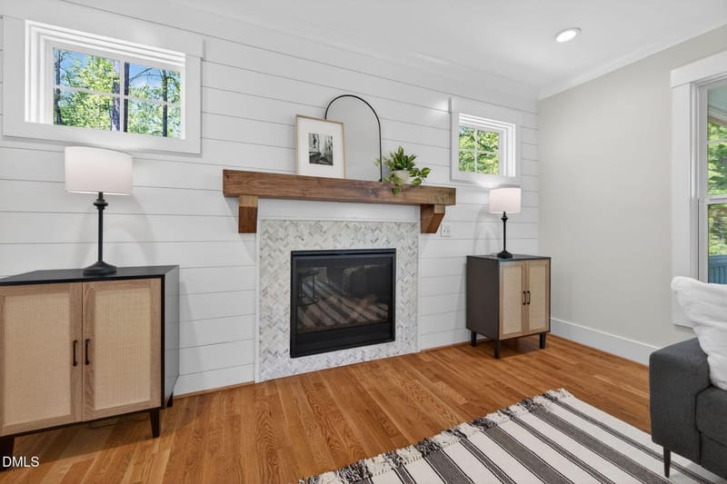 Living room with white shiplap walls, wooden mantel fireplace with herringbone tile surround, black insert, hardwood floors, and two windows.