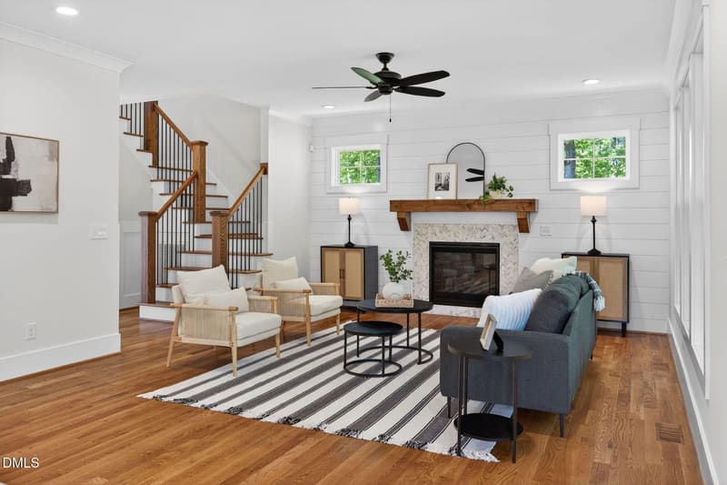 Bright living room with hardwood floors, striped rug, gray sofa, two armchairs, fireplace with wood mantel, and wooden staircase.