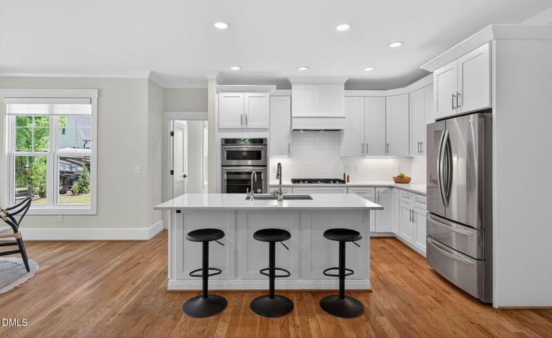 Bright modern kitchen with white shaker cabinets, large central island with three black stools, stainless steel fridge and double oven, hardwood floors.