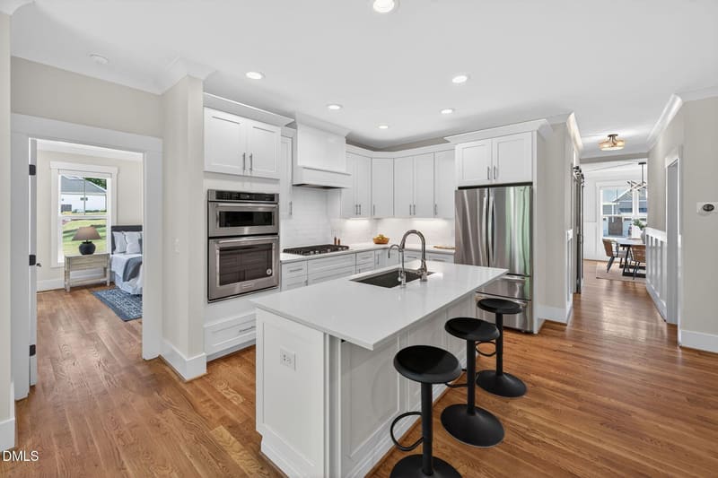 Modern white kitchen with quartz island, black bar stools, stainless steel appliances, and open views to bedroom and dining area.