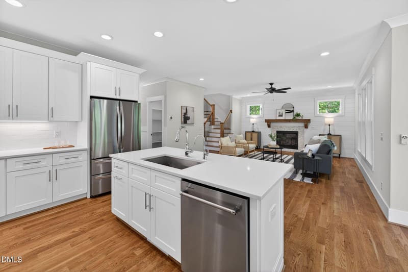 Bright open-concept kitchen with white cabinets, stainless steel appliances, large island with sink, and view to living room with fireplace and staircase.