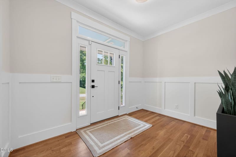 Bright foyer with white front door and transom, hardwood floors, wainscoting, area rug, and potted plant.