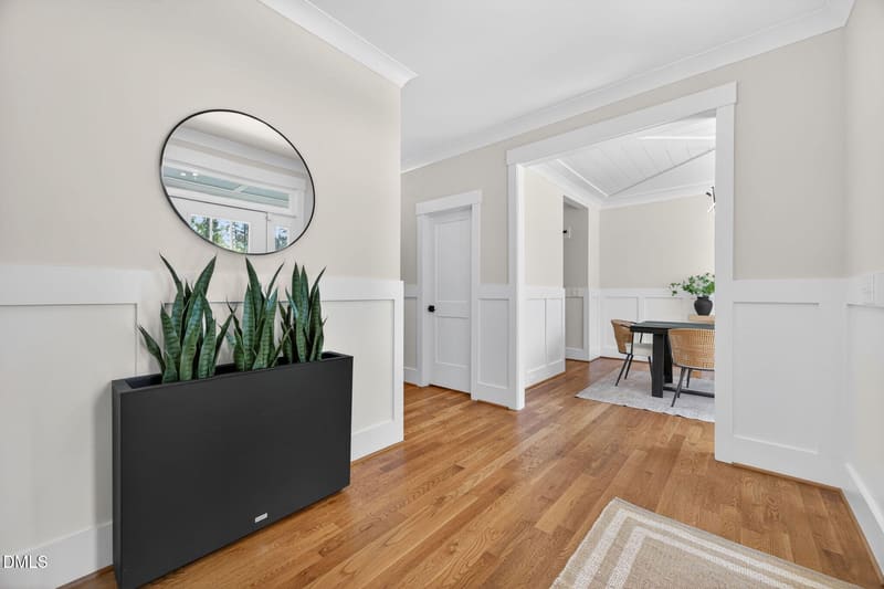 Bright entryway with hardwood floors, white wainscoting, round mirror above black planter with snake plants, open to dining area.