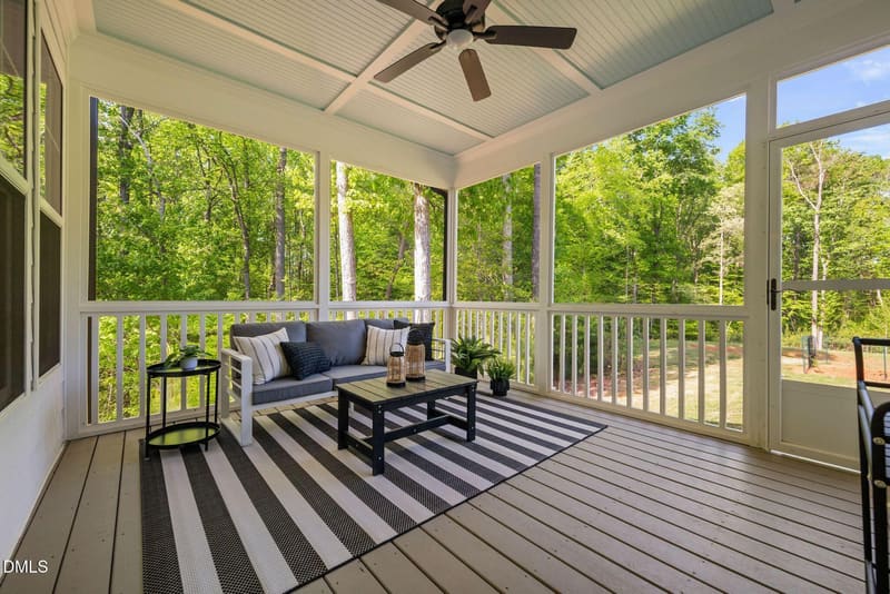 Screened porch with white trim, gray sofa with pillows, black coffee table, striped rug, ceiling fan, and large windows overlooking dense green forest.