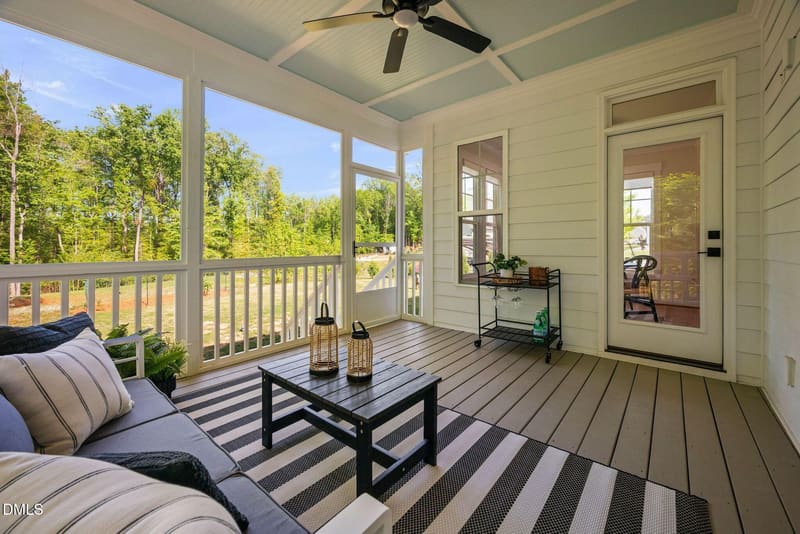 Screened porch with white walls, light blue ceiling fan, wood floors, striped rug, coffee table, and large windows overlooking lush forest.
