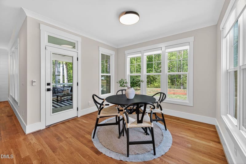 Dining room with round black table, four black-and-woven chairs on a circular rug, large windows overlooking trees, white door to deck, and hardwood floors.