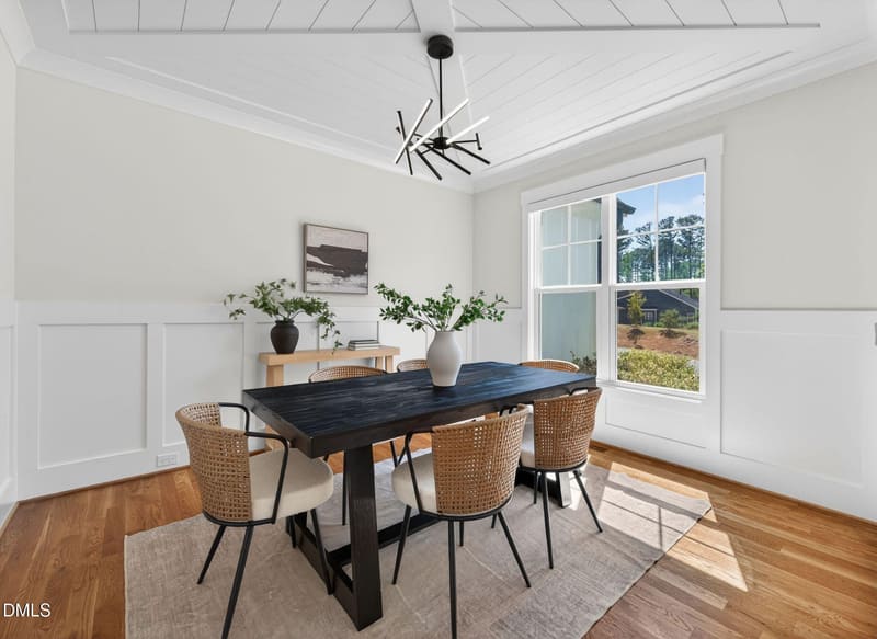 Dining room with dark wood table, four woven rattan chairs, white wainscoting, large window with natural light, modern chandelier, and abstract art.