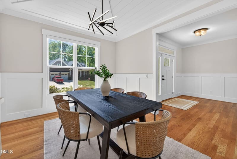 Bright dining room with dark wood table, four woven rattan chairs, modern black chandelier, large white-trimmed windows, and white wainscoting on beige walls.