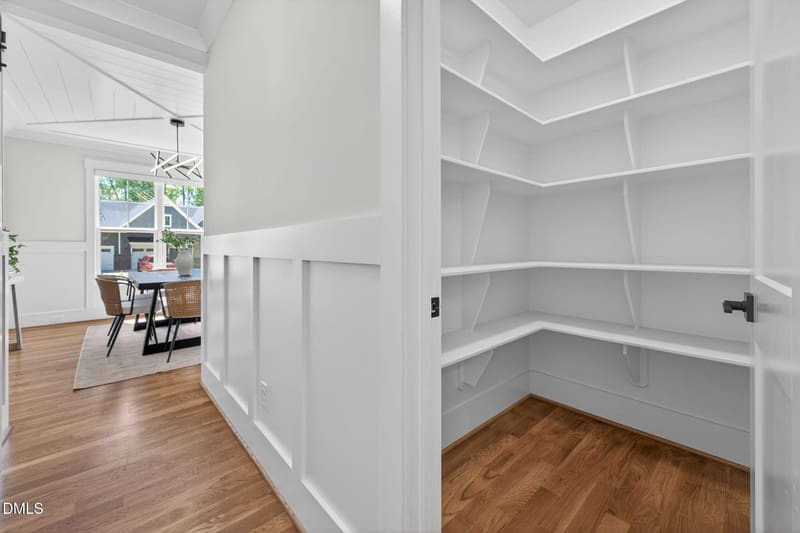 Open white door to empty walk-in closet with white built-in shelves, wood floor, and adjacent bright dining room with table, chairs, and large windows.