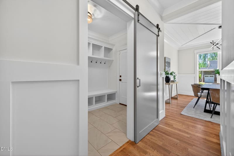 White sliding barn door partially open to mudroom with built-in white cubbies, hooks and bench on beige tile, adjacent to hardwood dining area with table and chairs.