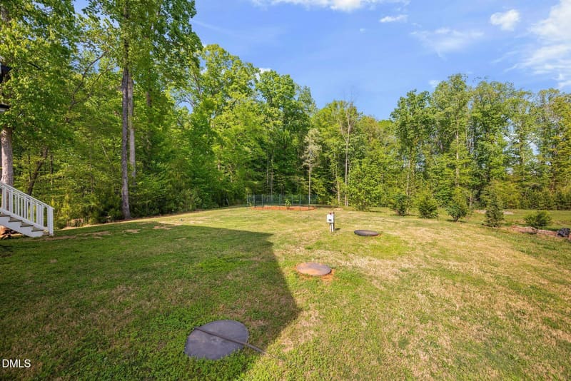 Large grassy backyard with utility covers, white stairs on left, and dense green forest under blue sky.