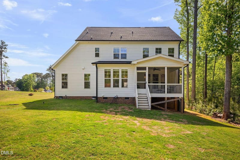 Backyard view of a two-story white house with screened porch, stairs, multiple windows, and brick foundation on a green lawn with trees.