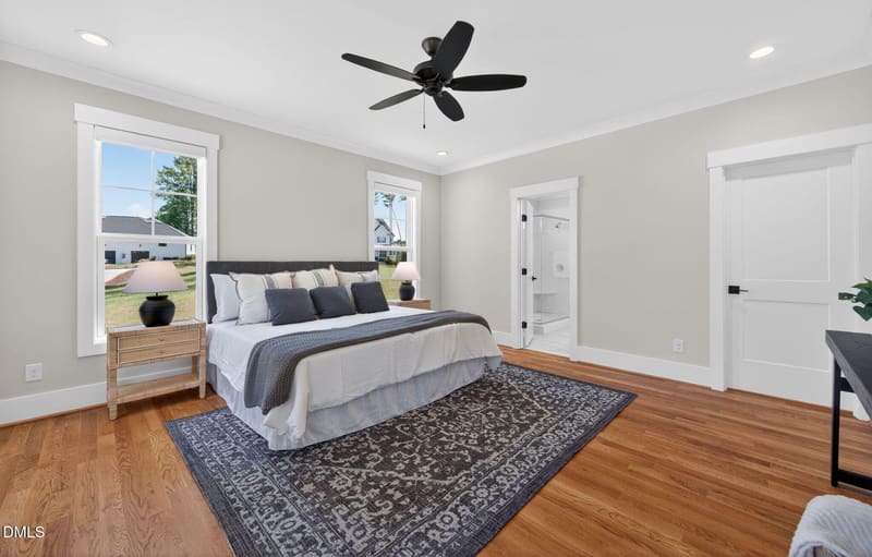 Spacious bedroom with king bed in white and gray linens, dark hardwood floors, patterned rug, black ceiling fan, and open door to en-suite bath.