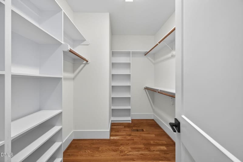 Empty walk-in closet with white built-in shelves, wooden hanging rods, white walls, and hardwood flooring.