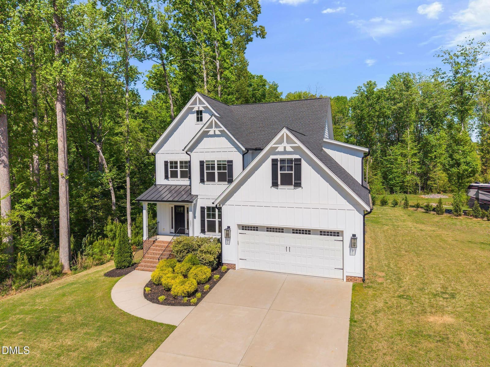 Two-story white house with dark gray roof, black shutters, attached garage, concrete driveway, and landscaped yard surrounded by tall green trees.
