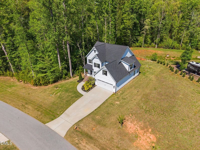 Aerial view of a two-story white house with dark roof, attached garage, concrete driveway, large lawn, and dense forest backdrop with an RV parked nearby.