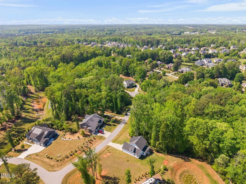 Aerial view of a suburban neighborhood with houses featuring dark roofs, surrounded by dense green trees and winding roads under a clear blue sky.